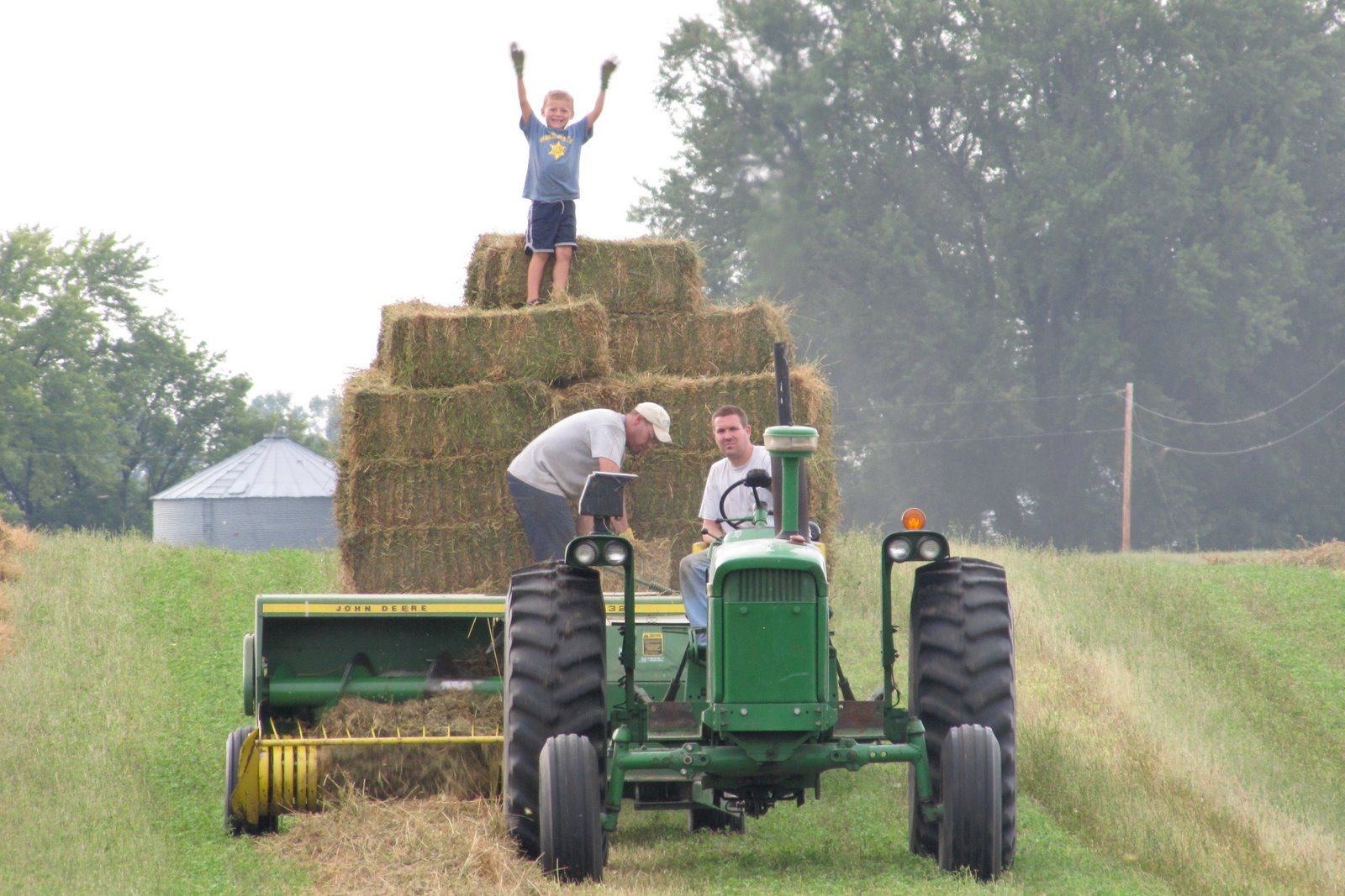 Free-range childhood: a young boy cheering on top of stacked hay bales while two men work a green John Deere tractor and baler