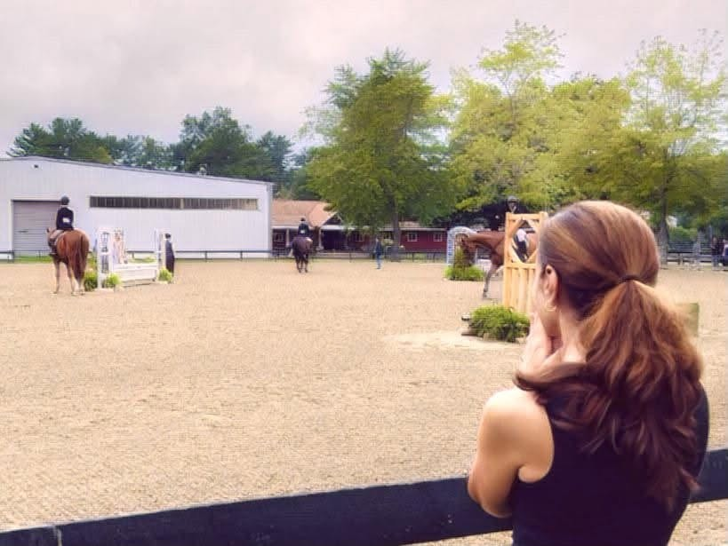 JoAnn watching a horse show ring from the rail as riders and horses warm up on the footing