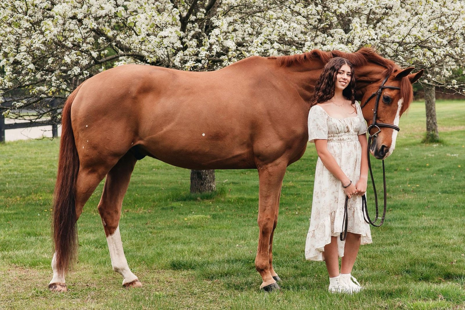 Piper standing with her chestnut horse under blossoming pear trees, the horse resting its head against her shoulder